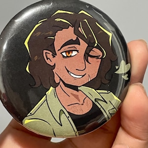 May include: A round button with an illustration of a smiling person with brown hair and a tan complexion. The person is wearing a green shirt over a black shirt. The background is black.