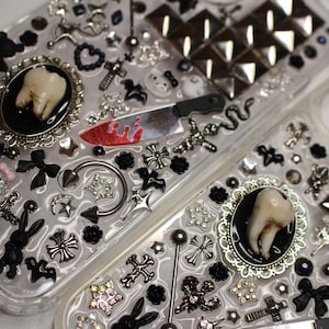 May include: Two clear phone cases decorated with black and silver charms, including a tooth, a knife, crosses, stars, and a snake. The cases are covered in rhinestones and have a studded design.