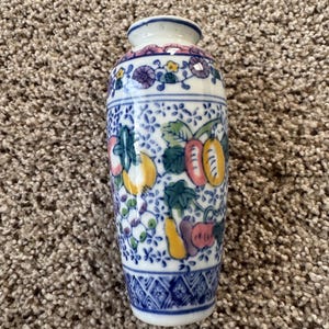 Puede incluir: Un pequeño jarrón de cerámica con base blanca que presenta un patrón pintado a mano de frutas y flores en azul, amarillo, rosa y verde. El diseño incluye un borde geométrico azul y blanco en la base y una banda floral alrededor del cuello.