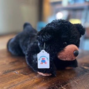 May include: Black plush toy bear lying on a wooden surface. The bear has a brown nose and a white tag that says "Mr. Poppers Pals".