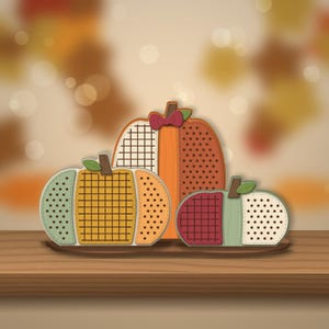 Può includere: Un set decorativo di tre ornamenti piatti a forma di zucca disposti su una base. Le zucche presentano motivi vari tra cui griglie, punti e colori solidi nelle tonalità arancione, giallo, verde, rosso e crema, con piccoli steli e foglie marroni.
