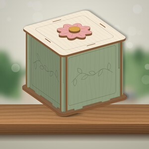 May include: A square wooden box with a light-colored lid featuring a pink flower with a yellow center. The sides of the box are sage green with brown edges and feature a delicate engraved vine and leaf pattern on the visible exterior panels.