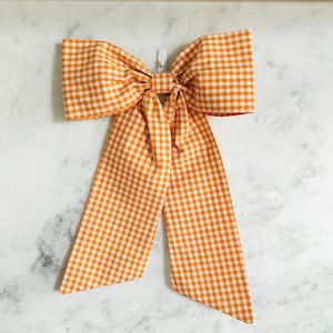 May include: A large fabric bow featuring an orange and white gingham check pattern. The bow has two wide, structured loops at the top and two long, rectangular tails hanging downwards, with a small white plastic clip visible at the center.