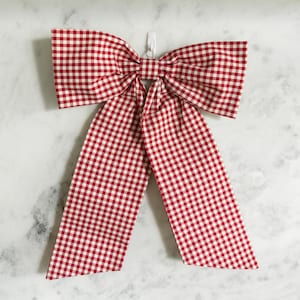 May include: A large decorative bow featuring a classic red and white gingham check pattern. The bow consists of two wide, horizontal loops gathered at the center and two long, vertical fabric tails that extend downward from the central knot.