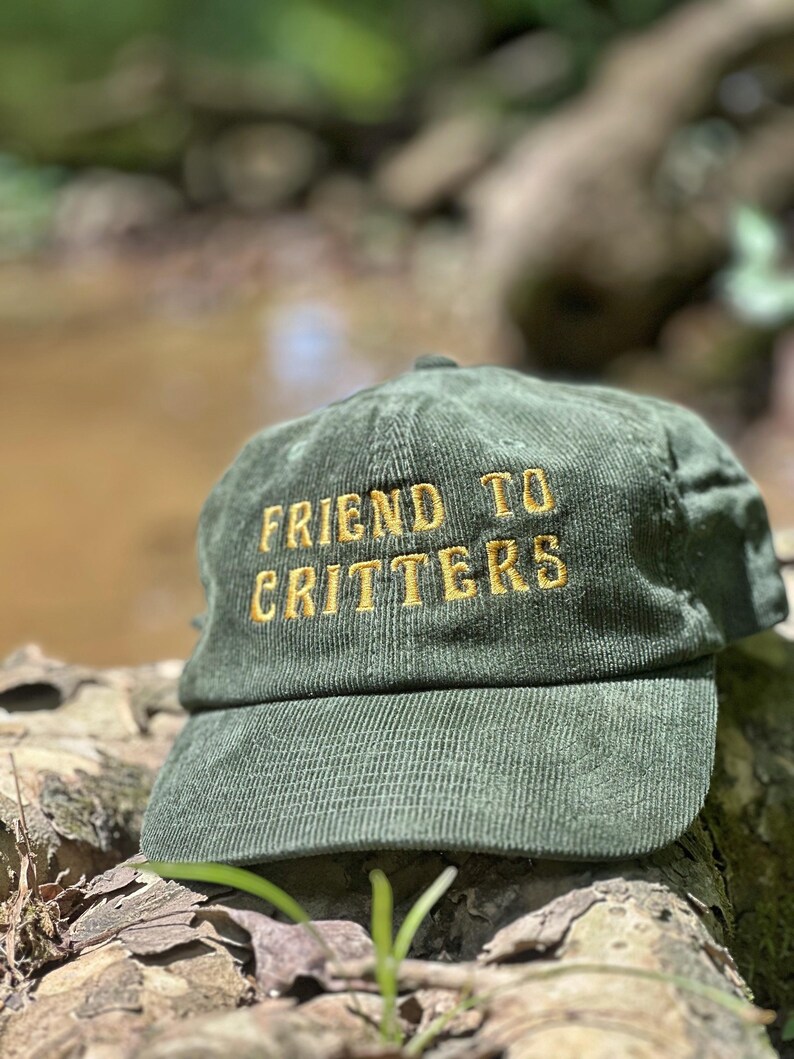 May include: An olive green corduroy baseball cap featuring the embroidered text "FRIEND TO CRITTERS" in gold thread across the front panels. The hat has a structured crown, a curved brim, and a classic six-panel design with visible vertical texture.