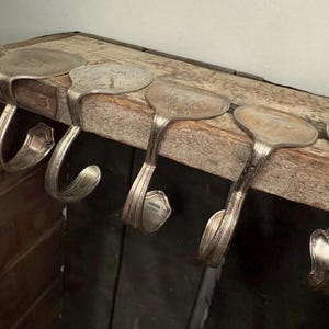 Könnte beinhalten: Fünf silberne Löffelhaken, montiert auf einem verwitterten Holzbrett. Die Löffel sind zu Haken gebogen, was ein rustikales, upgecyceltes Design zeigt. Die Haken sind an einer dunklen Holzoberfläche befestigt.