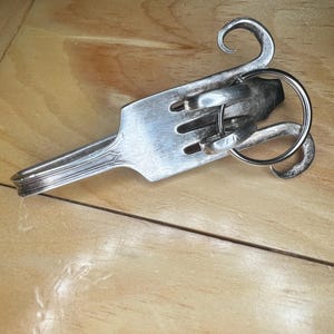 May include: A handcrafted silver fork transformed into a unique keychain. The fork's tines are intact, with decorative swirls added to the handle and a key ring attached. The piece is displayed on a wooden surface.