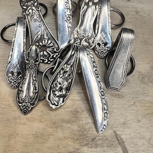 May include: A selection of vintage silver spoon keyrings. Each keyring showcases a spoon head with intricate floral and scrollwork patterns. The spoons are attached to black metal key rings, creating a unique and repurposed accessory.