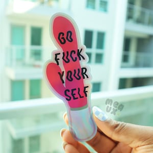 May include: A pink and red cactus-shaped sticker with the text "GO FUCK YOUR SELF" in black. The sticker has a holographic effect and is held up in front of a window.