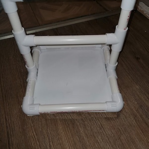 May include: A white PVC pipe frame with a black handle. The frame is assembled with 90-degree elbows and straight pipe sections. The frame is sitting on a wooden floor.