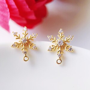 May include: Two gold-colored snowflake charms with clear crystal accents. Each charm has a loop at the bottom for attaching to jewelry. The snowflake design features six points with smaller crystals.