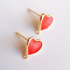 May include: A pair of gold heart-shaped earrings with a red enamel fill. The earrings have a post back closure.