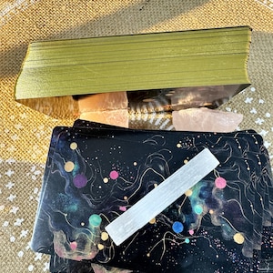 May include: A deck of tarot cards with a black background and colorful abstract designs. The cards are stacked on top of a white crystal and a pink crystal. The cards are surrounded by a beige fabric with a white pattern.