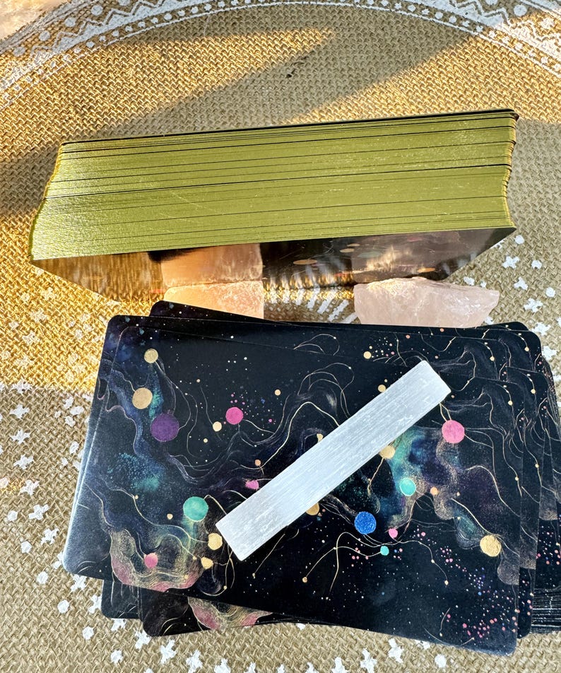 May include: A deck of tarot cards with a black background and colorful abstract designs. The cards are stacked on top of a white crystal and a pink crystal. The cards are surrounded by a beige fabric with a white pattern.