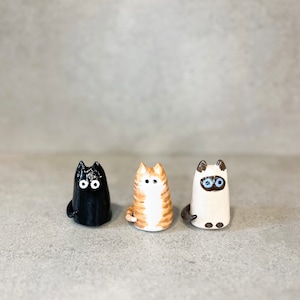 Puede incluir: Tres figuritas de cerámica de gatos, una negra, una naranja y blanca y una blanca con la cara gris. Las figuritas están todas de pie y tienen ojos pintados.