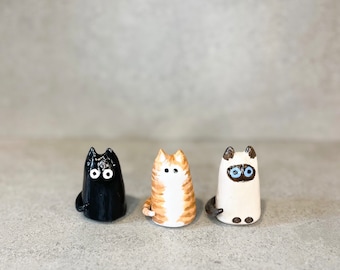 Puede incluir: Tres figuritas de cerámica de gatos, una negra, una naranja y blanca y una blanca con la cara gris. Las figuritas están todas de pie y tienen ojos pintados.