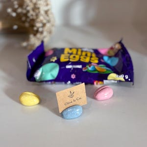 May include: A purple packet of Mini Eggs, with a 200 g weight, and three pastel-coloured egg-shaped candies. A small card holder with the text "Olive It Co." is also visible.