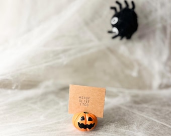 May include: A small orange pumpkin figurine with a black jack-o'-lantern face, holding a brown card with the text "@shop olive itco".