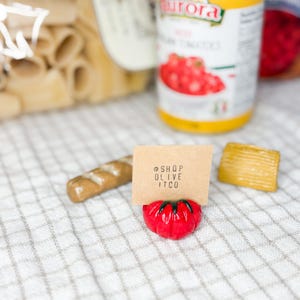 Peut inclure: Nature morte avec un ornement en forme de tomate rouge tenant une pancarte avec l'inscription "SHOP OLIVE ITCO". Une boîte de tomates en dés Aurora et un sac de pâtes sont visibles. Une petite baguette et un objet rectangulaire jaune sont également présents.