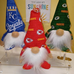 May include: Three decorative gnomes with white beards and fabric hats. One gnome has a blue hat with a baseball and "Royals" text, another has a red hat with a football and "Chiefs" text, and the third has a green hat with a football and "Packers" text.