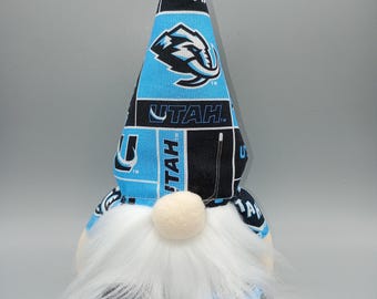 May include: A gnome doll with a long white beard and a cream-colored nose. The gnome's hat is patterned with black and light blue fabric, featuring a logo and the word "UTAH". The gnome has a light-colored face and a fluffy white beard.