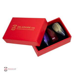 Könnte beinhalten: Eine leuchtend rote rechteckige Schachtel mit dem goldenen Aufdruck DELARAMM.COM Exclusive Musical Instrument auf dem Deckel. Im Inneren befinden sich drei tropfenförmige Plektren in Violett, Dunkelrot und Metallic-Silber in einer schwarzen Schaumstoffeinlage.