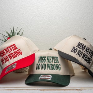 May include: Three baseball caps with the text "MISS NEVER DO NO WRONG" embroidered on the front. The caps have different color schemes: red and cream, green and cream, and black and cream. The caps are displayed on a white surface.