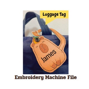 Könnte beinhalten: Ein braunes Leder-Gepäckanhänger mit einem Cartoon-Bären-Design und dem Namen "James" bestickt. Der Anhänger ist an einer blauen Tasche befestigt.