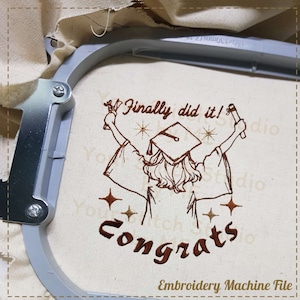 Pode incluir: Desenho bordado de um graduado com capelo e beca, levantando os braços no ar. O texto "Finally did it! Congrats" está bordado abaixo do graduado. O desenho está em um tecido branco.