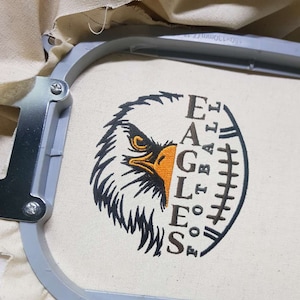 May include: Embroidered design of an eagle's head with the word "EAGLES" vertically down the side and "FOOTBALL" vertically down the other side. The design is on a white fabric.
