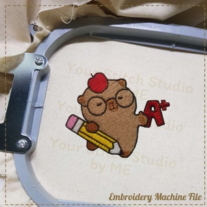 Puede incluir: Diseño bordado de un oso marrón con gafas y una manzana en la cabeza, sosteniendo un lápiz y una A+ roja. El diseño está en una tela blanca en un aro.
