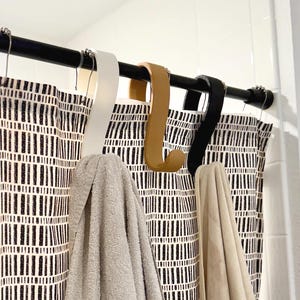 May include: A bathroom scene featuring a black shower rod with three S-shaped shower curtain hooks in white, tan, and black. Two towels in light gray and beige hang from the hooks. The shower curtain has a black and white geometric pattern.