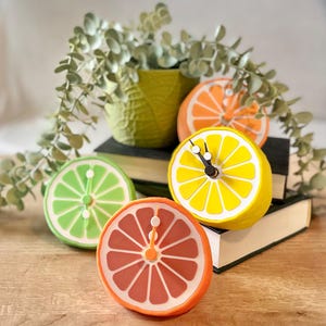 May include: A collection of fruit-shaped clocks, including a lime, orange, lemon, and grapefruit, arranged on a wooden surface. The clocks have black clock hands and are set against a backdrop of books and greenery. The clocks are a fun and colorful addition to any room.
