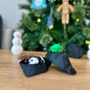 Op de afbeelding: Een zwart-witte panda, een bruine eekhoorn en een groen kikkerfiguurtje bevinden zich in een zwarte, open, hartvormige container. De container staat op een houten oppervlak.