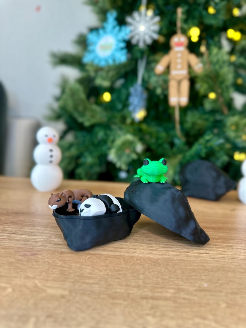 Op de afbeelding: Een zwart-witte panda, een bruine eekhoorn en een groen kikkerfiguurtje bevinden zich in een zwarte, open, hartvormige container. De container staat op een houten oppervlak.