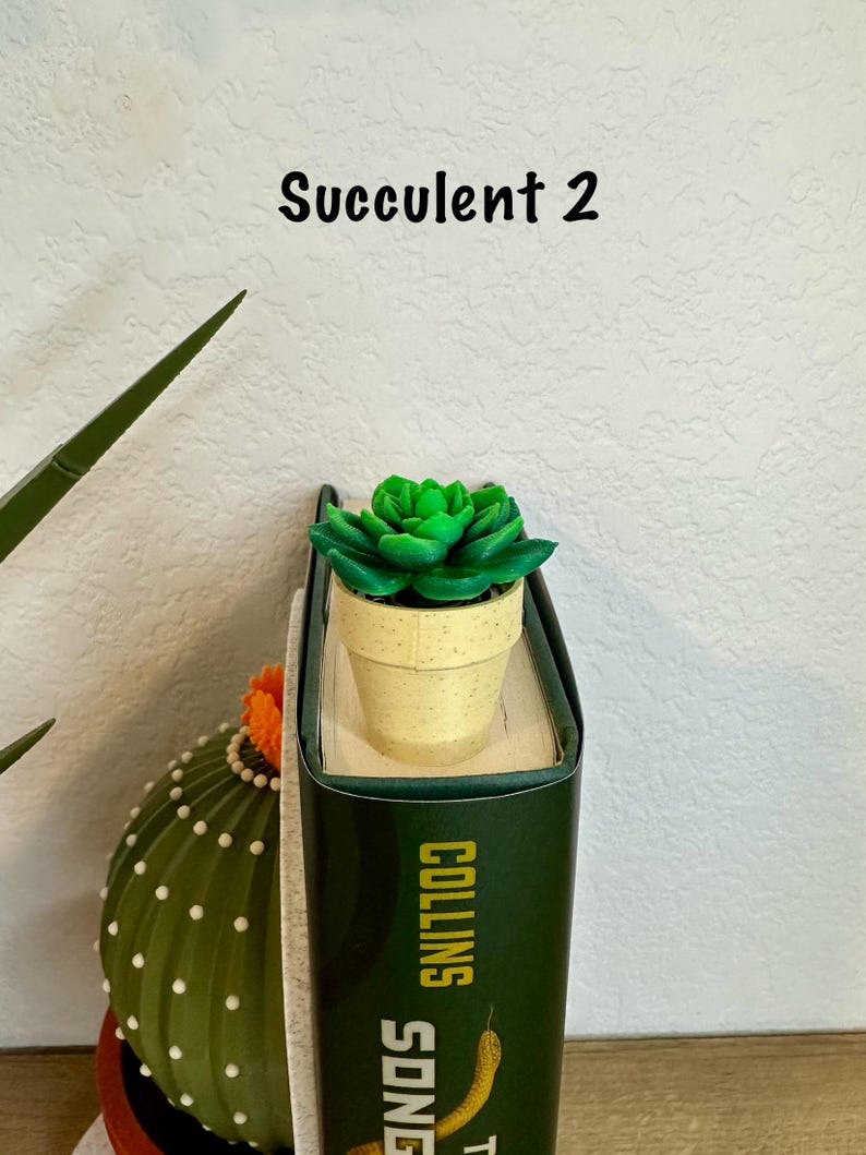 May include: A small green succulent plant in a beige pot, used as a bookmark on a green book with the text "COLLINS SONG" on the spine. The text "Succulent 2" is written above the plant.