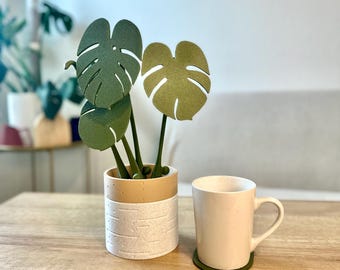 May include: A white ceramic planter with a brown base holds a faux green monstera plant. The plant has three leaves, two green and one light green. A white ceramic mug with a green coaster sits on a wooden table.