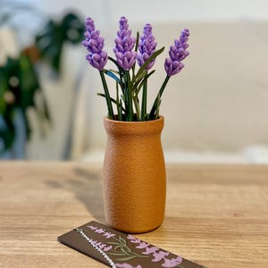 May include: A small, terracotta-colored vase holds several purple lavender stems with green leaves. A decorative bookmark with a lavender design and a green tassel lies on a wooden surface, complementing the floral arrangement.