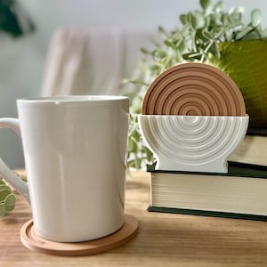 May include: A white ceramic mug sits on a wooden coaster. A set of brown and white coasters are in a white holder. The coasters have a concentric circle design. The items are on a wooden surface.