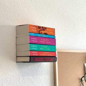 May include: A stack of hardcover books with colorful spines, including titles like "A Court of Silver Flames" and "A Court of Thorns and Roses" by Sarah J. Maas. The books are arranged on a shelf against a white wall.