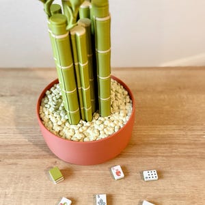 May include: A decorative plant sculpture featuring multiple green bamboo-like stalks with segmented details, potted in a round terracotta-colored container filled with small white pebbles, accompanied by seven small rectangular game tiles.