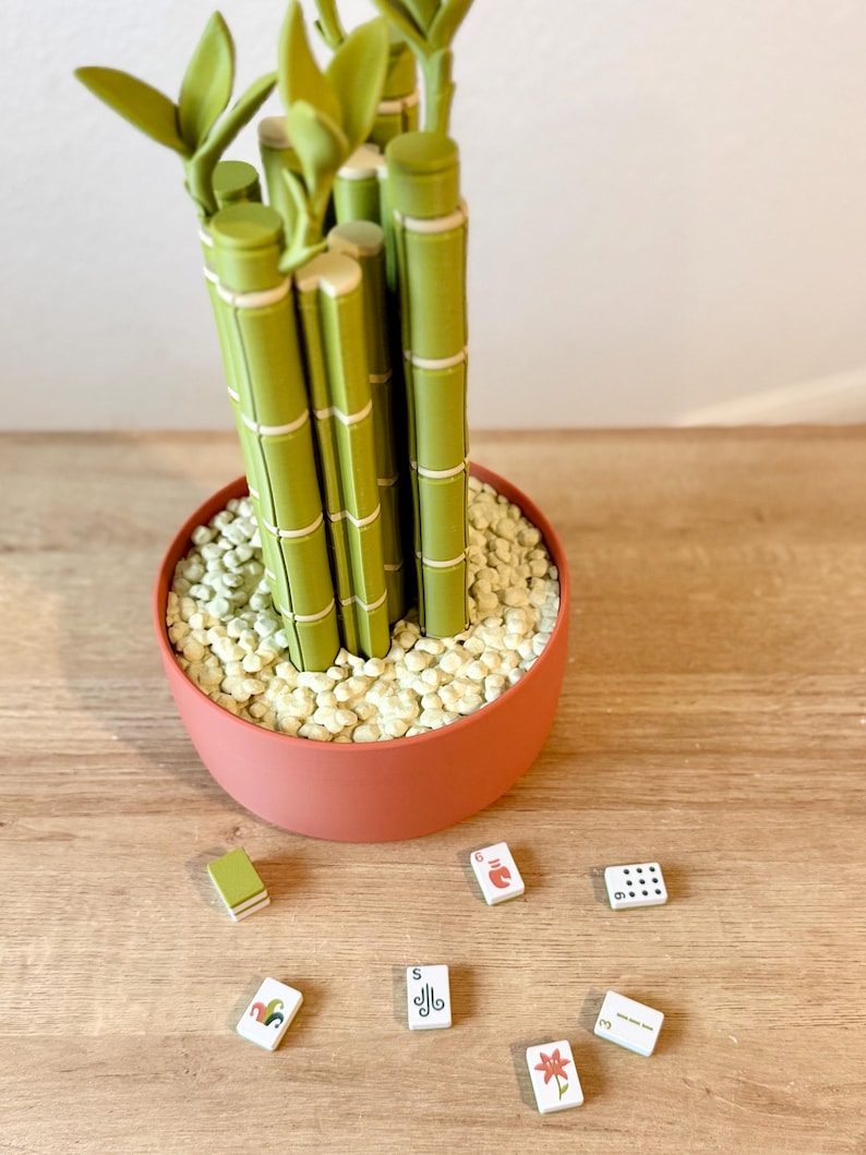 May include: A decorative plant sculpture featuring multiple green bamboo-like stalks with segmented details, potted in a round terracotta-colored container filled with small white pebbles, accompanied by seven small rectangular game tiles.