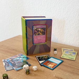 May include: A rectangular, book-shaped storage case with a rainbow-colored frame and a central window displaying an Alakazam trading card. Several loose trading cards and small, numbered gaming dice are scattered around the base of the case.