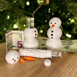 May include: Two white snowman-shaped money banks with black eyes and buttons. One snowman has a carrot nose and the other has a rolled up dollar bill inside. There is a white plastic tube with a carrot nose and a $5 bill on the table.
