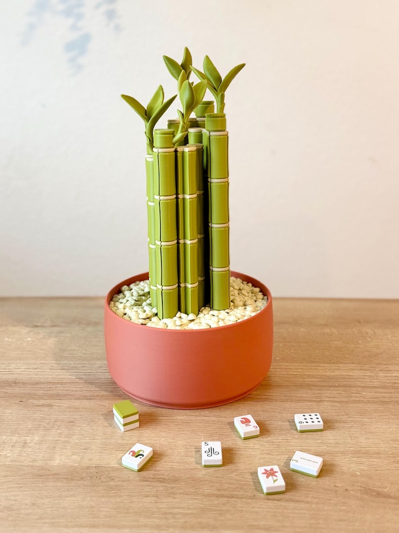 May include: A decorative bamboo plant sculpture constructed from green plastic segments, potted in a round terracotta-colored container filled with small white stones. Six small rectangular tiles with various printed symbols are scattered nearby.