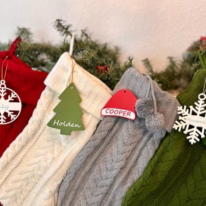 May include: Four Christmas stockings with personalized ornaments. The stockings are red, white, gray, and green. The ornaments are a snowflake with the name "Lily", a Christmas tree with the name "Holden", a Santa hat with the name "Cooper", and a snowflake with the name "Isabelle".