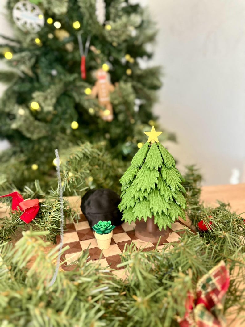 May include: A miniature green Christmas tree with a yellow star sits on a brown and beige chessboard next to a small potted succulent and a black hat. The scene is surrounded by green Christmas tree branches.