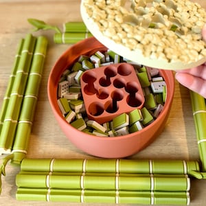 May include: A round terracotta-colored container filled with small green and white game tiles and a central square insert with nine circular holes. The container has a textured, light-colored lid and is surrounded by four green bamboo-shaped pieces.