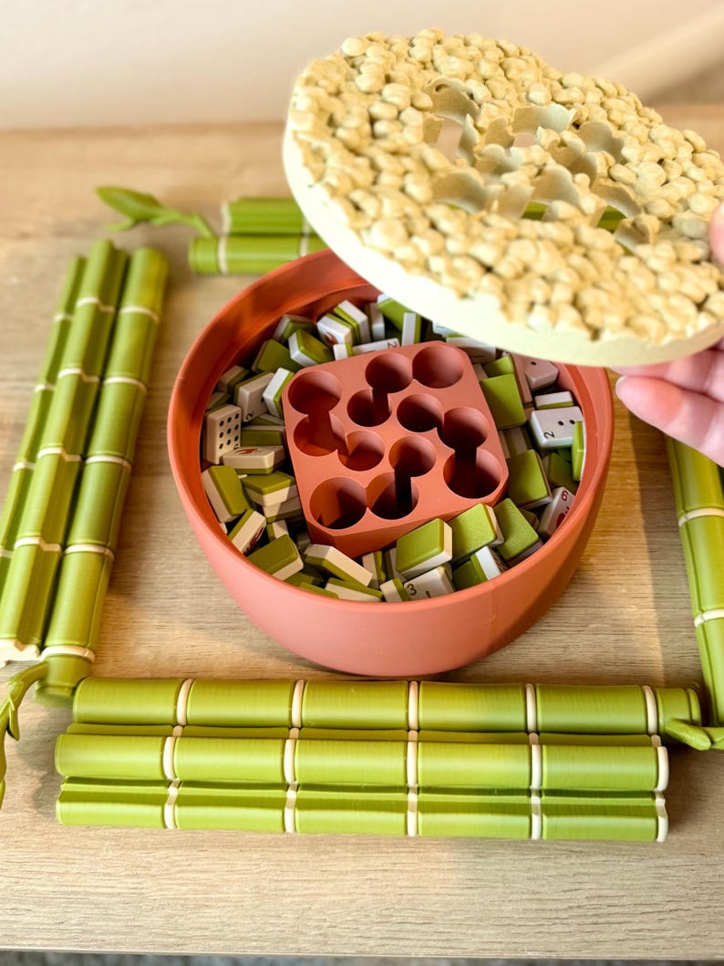 May include: A round terracotta-colored container filled with small green and white game tiles and a central square insert with nine circular holes. The container has a textured, light-colored lid and is surrounded by four green bamboo-shaped pieces.