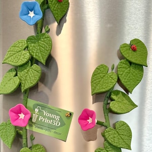 May include: A 3D printed green vine with leaves and flowers. The vine has pink, blue, and white flowers. There are also red ladybugs on the vine. The vine is attached to a metal surface. The text "Young Print3D" is on a white card with a green border. The text "ints - plants - pots - decor" is below the company name.
